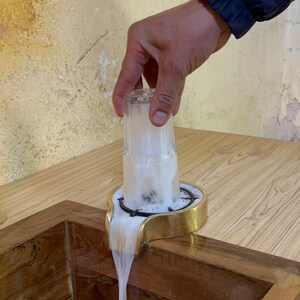 May include: A glass of foamy liquid is being poured into a wooden sink. The glass is being held by a hand and is resting on a gold-colored metal stand. The liquid is flowing out of the glass and into the sink.