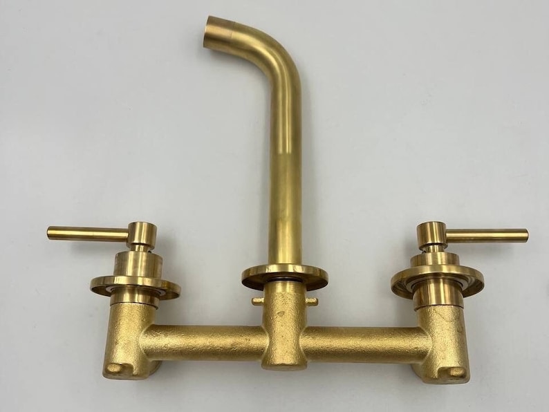 Op de afbeelding: Een gouden kraan met twee handvatten en een uitloop. De kraan is ontworpen voor een keukengoten.