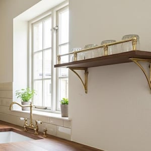 May include: A wall-mounted wooden shelf featuring a polished brass railing and two matching brass support brackets. The shelf holds four clear glass tumblers arranged in a row, providing a functional storage solution for a kitchen space.