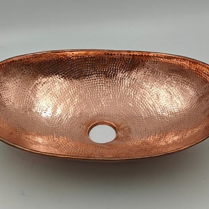 May include: Oval copper sink with a hammered texture and a drain hole. The sink has a polished finish, reflecting light. The copper color is a warm, reddish-brown. This sink is designed for bathroom use.