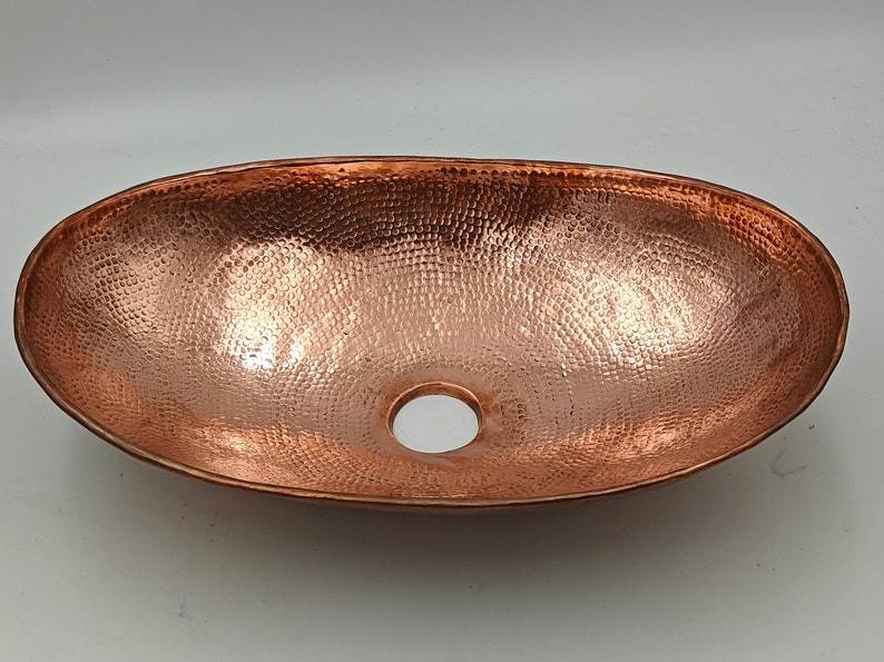 May include: Oval copper sink with a hammered texture and a drain hole. The sink has a polished finish, reflecting light. The copper color is a warm, reddish-brown. This sink is designed for bathroom use.