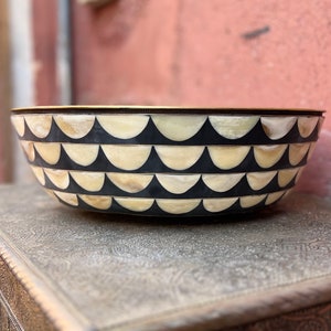 May include: Decorative bowl with a black and cream scallop pattern. The bowl has a gold rim and sits on a silver surface. The bowl's design is repeated in horizontal rows around the exterior.