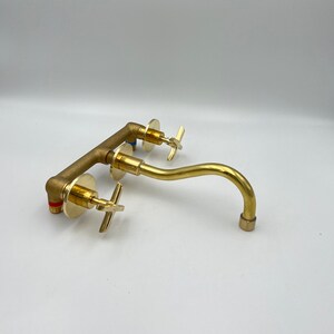 May include: A gold-toned wall-mounted faucet with a curved spout and two cross-shaped handles.