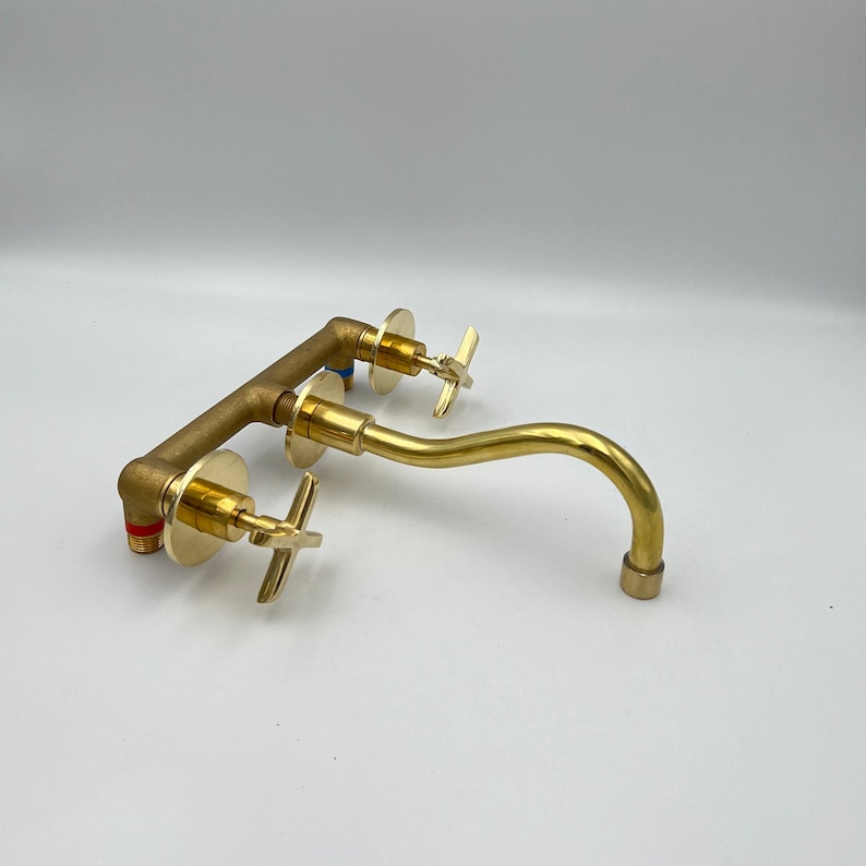 May include: A gold-toned wall-mounted faucet with a curved spout and two cross-shaped handles.