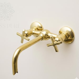 May include: A gold-colored wall-mounted faucet with cross-shaped handles. The faucet has a curved spout and is made of a shiny, metallic material. The fixture is mounted on a white wall.