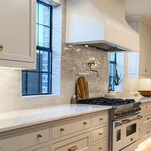 May include: A white kitchen with white cabinets, a white range hood, and white subway tile backsplash. The kitchen has a stainless steel range with a black cooktop and a white oven. The kitchen also has a white countertop and gold cabinet hardware.
