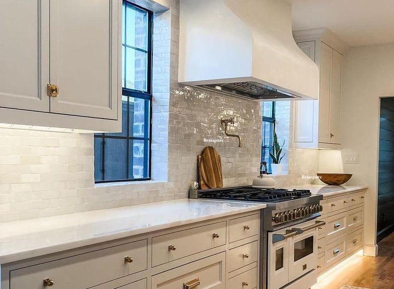 May include: A white kitchen with white cabinets, a white range hood, and white subway tile backsplash. The kitchen has a stainless steel range with a black cooktop and a white oven. The kitchen also has a white countertop and gold cabinet hardware.