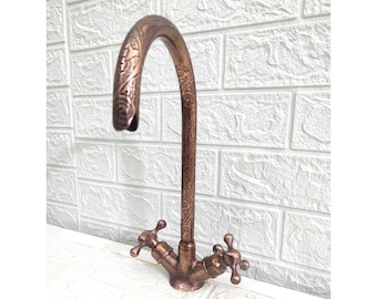 May include: A copper kitchen faucet with an ornate design and two handles. The faucet has a high arc spout and is mounted on a white countertop.