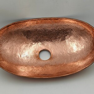 May include: Oval copper sink with a hammered texture and a central drain hole. The sink has a polished finish, reflecting light and showcasing the warm tones of the copper. A decorative and functional bathroom fixture.