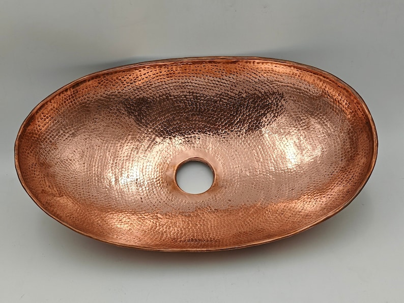 May include: Oval copper sink with a hammered texture and a central drain hole. The sink has a polished finish, reflecting light and showcasing the warm tones of the copper. A decorative and functional bathroom fixture.