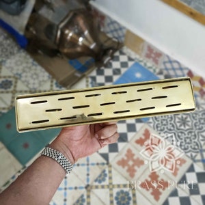 May include: A gold-colored brass drain cover with a rectangular shape and a slotted design. The cover has multiple parallel slots running the length of the drain cover.