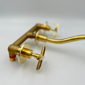 May include: A close-up of a gold-toned faucet with three separate spouts and handles. The faucet has a modern design with a sleek, curved shape. The handles are cross-shaped and are made of the same gold-toned metal.