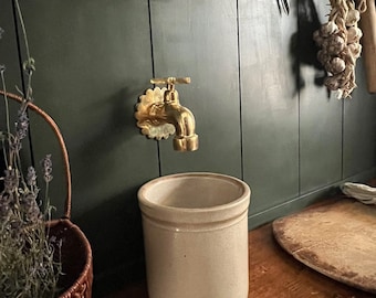 May include: A brass faucet with a decorative design mounted on a dark green wall. A beige ceramic crock sits on a wooden countertop.