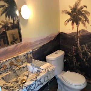 Op de afbeelding: Een wit toilet met een wit toiletbril in een badkamer met een marmeren wastafel en een tropische behangmuur met palmbomen en bergen.