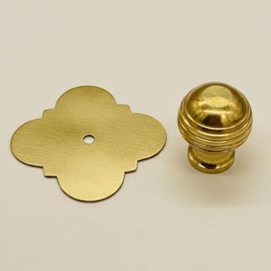 Può includere: Un set di due elementi hardware in ottone. Uno è un pomello decorativo con scanalature orizzontali e una parte superiore arrotondata. L'altro è una piastra posteriore a quattro lobi con un singolo foro. Entrambi hanno una finitura dorata lucida.