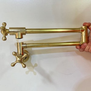May include: A gold-toned kitchen tap with a swivel spout and two cross-shaped handles. The tap is mounted on a wall.