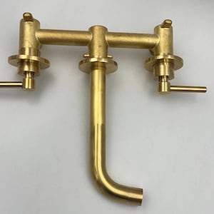 Op de afbeelding: Een gouden messing kraan met twee handvatten en een uitloop. De kraan is ontworpen voor wandmontage en heeft een moderne, minimalistische stijl.
