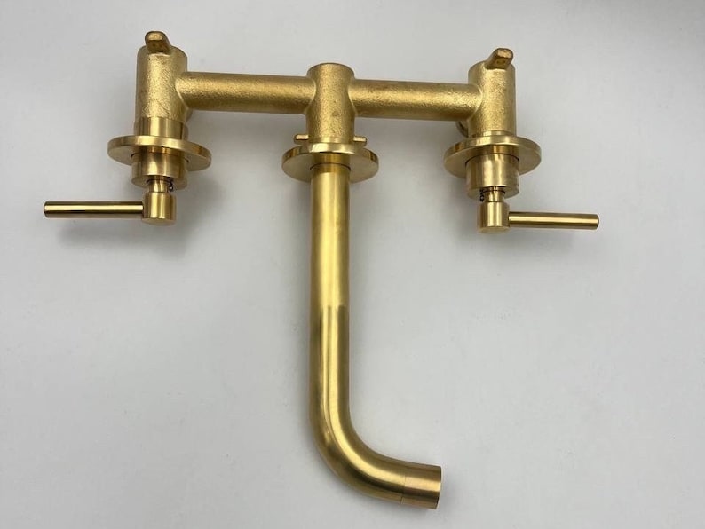 Op de afbeelding: Een gouden messing kraan met twee handvatten en een uitloop. De kraan is ontworpen voor wandmontage en heeft een moderne, minimalistische stijl.