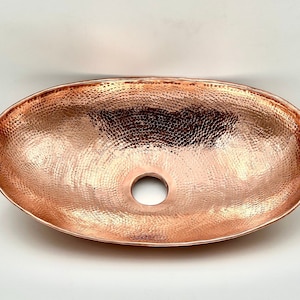May include: Oval copper sink with a hammered texture. The sink has a polished finish and a drain hole in the center. The copper sink is a bathroom fixture.