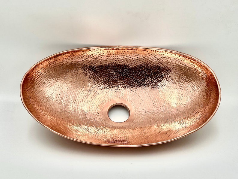May include: Oval copper sink with a hammered texture. The sink has a polished finish and a drain hole in the center. The copper sink is a bathroom fixture.