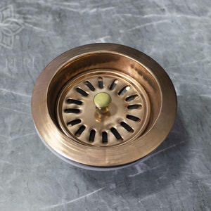 May include: A copper sink strainer with a brass drain stopper. The strainer has a circular design with multiple holes.