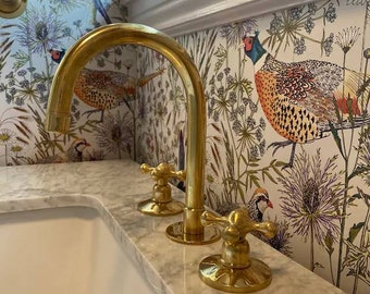 Könnte beinhalten: Goldenes Badezimmerarmatur mit kreuzförmigen Griffen, montiert auf einer weißen Marmor-Arbeitsplatte. Die Armatur befindet sich vor einem weißen Hintergrund mit einem floralen Muster mit Vögeln.
