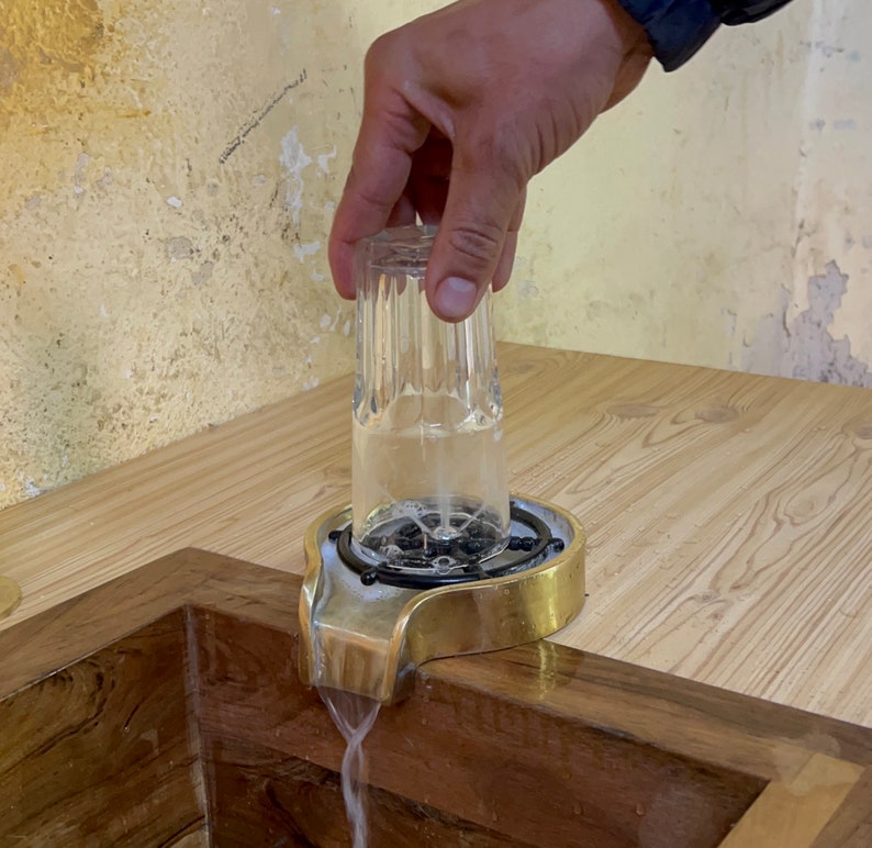May include: A glass is being placed over a brass water fountain spout with a black metal ring. Water is flowing from the spout into the glass.