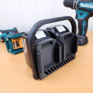 May include: Black battery holder for Makita power tools, with a handle and slots for two batteries. A Makita drill and impact driver are in the background, all on a wooden surface. The holder is designed to carry and store batteries.