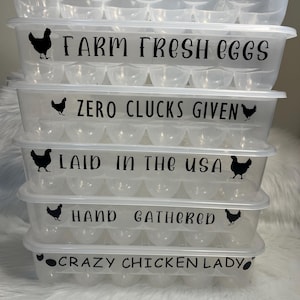 May include: A clear plastic egg carton with five rows of egg holders. Each row has a black and white sticker with a funny saying about farm fresh eggs. The sayings are "Straight Outta The Coop", "Farm Fresh Eggs", "Zero Clucks Given", "Laid In The USA", "Hand Gathered", and "Crazy Chicken Lady".