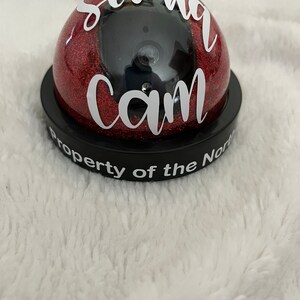 May include: A red glitter dome with a black camera inside and the words "Santa Cam" and "Property of the North Pole" written in white.