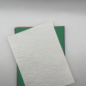 May include: White cardstock with an embossed pattern of graduation caps. The cardstock is layered on top of green, blue, and pink cardstock.