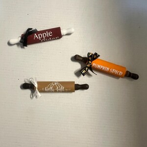 May include: Three decorative rolling pins with text and bows. One is burgundy with "Apple Cider" text, another is orange with "Pumpkin Spice" text, and the third is tan with "hello fall" text and a pumpkin graphic.