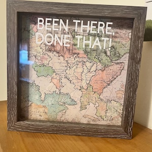 May include: A framed vintage map with the text "BEEN THERE, DONE THAT!" in white. The map features a variety of colors, including green, pink, and beige. The frame is a dark, weathered gray.