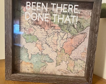 May include: A framed vintage map with the text "BEEN THERE, DONE THAT!" in white. The map features a variety of colors, including green, pink, and beige. The frame is a dark, weathered gray.
