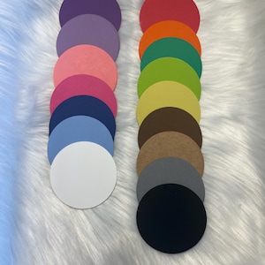 May include: Two columns of colorful, round paper cutouts. The left column features shades of purple, pink, blue, and white. The right column displays red, orange, green, yellow, brown, gray, and black circles. The cutouts are arranged on a white, fluffy surface.