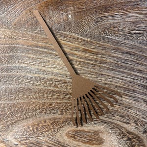 May include: A brown paper cutout of a garden rake. The rake has a long, thin handle and a fan of tines at the end. The cutout is placed on a wooden surface with a natural wood grain pattern.
