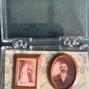 May include: Miniature framed photographs, one rectangular and one oval, presented in a clear plastic case. The rectangular frame contains a wedding photo, and the oval frame shows a man's portrait. The background is patterned paper.