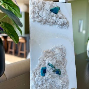 Op de afbeelding: Wit canvas met een getextureerd, besneeuwd oppervlak en blauwe agaatstenen. Het canvas is rechtsonder getekend met "Camilla Jewelry".