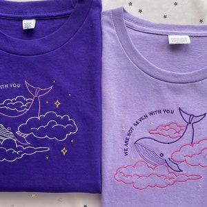 May include: Two purple t-shirts with a whale and clouds design. The text on the shirts reads "We are not seven with you".