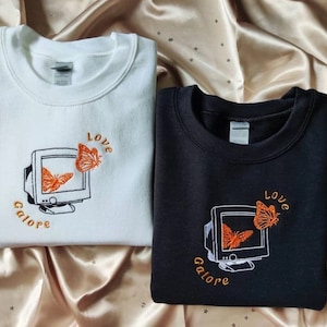 May include: Two sweatshirts with embroidered designs. One is white with orange butterflies and the text "Love Galore" and the other is black with orange butterflies and the text "Love Galore".