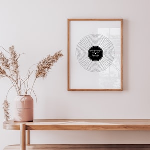 May include: A framed print featuring a circular design resembling a vinyl record. The design is composed of song lyrics in a circular pattern around a black center. The frame is a warm brown color, and the print is displayed on a white wall.