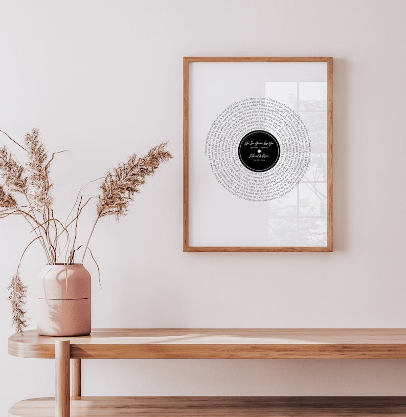 May include: A framed print featuring a circular design resembling a vinyl record. The design is composed of song lyrics in a circular pattern around a black center. The frame is a warm brown color, and the print is displayed on a white wall.