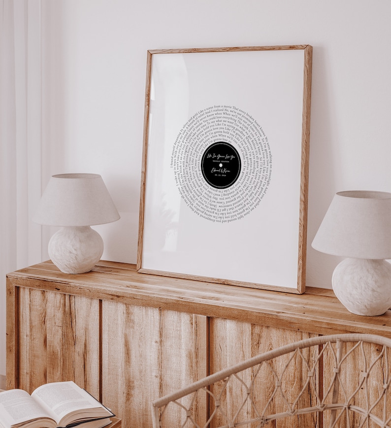 May include: A framed print featuring a circular design resembling a vinyl record. The design is composed of song lyrics in a circular pattern around a black center with text. The frame is a light brown color.