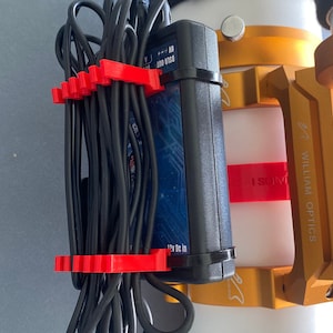 Puede incluir: Un adaptador de corriente negro con un diseño azul y plateado está unido a un soporte de metal dorado. Se utilizan bridas de cable rojas para organizar los cables negros.