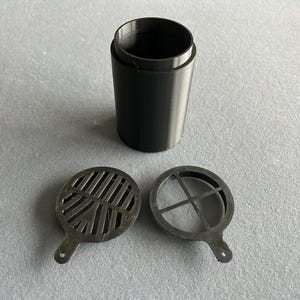 Pode incluir: Um recipiente cilíndrico preto com duas inserções redondas e ranhuradas correspondentes. O recipiente é aberto no topo e as inserções têm pequenas alças. Os itens estão em uma superfície cinza.