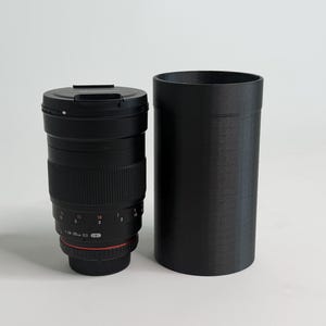 May include: A black camera lens with a red accent ring and a matching black cylindrical case. The lens has a ribbed grip and markings indicating focal length. The case is tall and cylindrical.