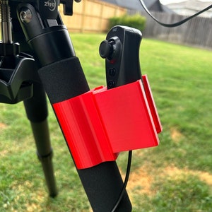May include: A red 3D printed holder for a black video game controller attached to a black tripod leg.