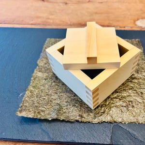 Może przedstawiać: Drewniane pudełko z pokrywką, leżące na arkuszu suszonych wodorostów. Pudełko wykonane jest z jasnego drewna i ma prosty design. Pokrywka ma mały uchwyt.