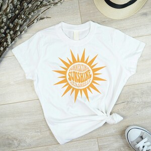 以下が含まれることがあります： 「Summertime Sunshine Seashells」の文字が入ったオレンジ色の太陽のグラフィックが特徴の白いTシャツ。Tシャツはウエストで結ばれています。麦わら帽子とスニーカーが背景に見えます。