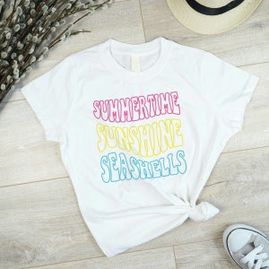 以下が含まれることがあります： ピンク、黄色、青で「SUMMERTIME SUNSHINE SEASHELLS」と書かれた白いTシャツ。シャツはウエストで結ばれています。麦わら帽子と白いスニーカーが背景に見えます。